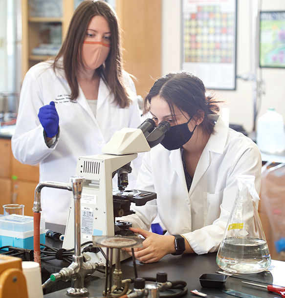 two female MSJ students in lab coats looking through microscope in classroom.