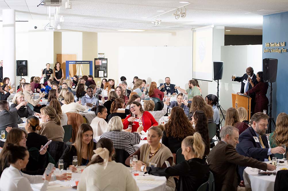 group of attendees at event at banquet tables listening to president speech at podium